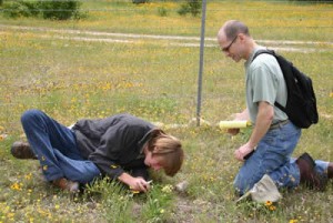 Two men kneeling in a field with a notebook and a magnifying glass.