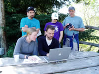 Three people standing and two sitting at a picnic table reviewing data on a laptop.
