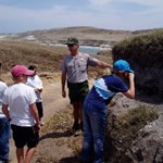 A man standing next to a rock outcropping explaining the geology to a group of kids.