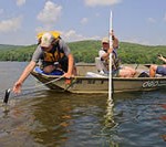 Man leaning over front of a boat in the water with an instrument to collect data.