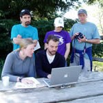 Three people standing and two sitting at a picnic table reviewing data on a laptop.